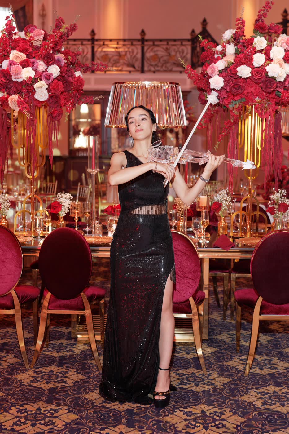 Sadie Varius, bow raised, the transparent violin in hand amid crimson roses and a crystal lampshade at The Legacy Castle